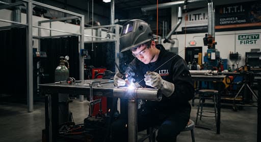 A striking industrial photography shot capturing a welding student in full protective gear meticulously performing a TIG weld on a steel joint